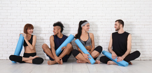 Group of sporty people with yoga mats sitting near white brick wall