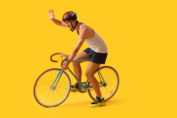 Young male cyclist riding bicycle on yellow background and waving hand