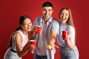 Young friends with cups and ball for beer pong on red background