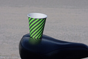 one paper black green striped paper cup with coffee stands on a leather bicycle seat on a gray background on the street