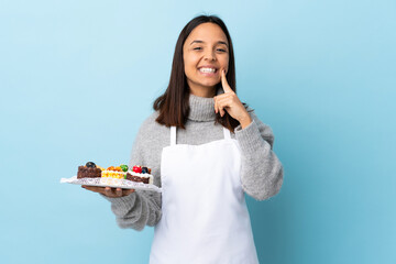 Pastry chef holding a big cake over isolated blue background smiling with a happy and pleasant...