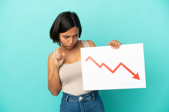 Young mixed race woman isolated on blue background holding a sign with a decreasing statistics arrow symbol with fingers crossing