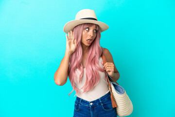 Young mixed race woman holding a beach bag with pamela isolated on blue background listening to...