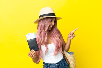 Young mixed race woman holding passport and beach bag isolated on yellow background pointing finger to the side and presenting a product