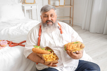 Overweight mature man with unhealthy food in bedroom. Overeating concept