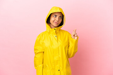 Young latin woman wearing a rainproof coat over isolated background with fingers crossing and...