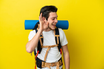 Young mountaineer Russian man with a big backpack isolated on yellow background listening to...
