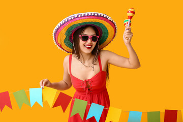 Happy young woman in Mexican sombrero hat, with maracas and garland on yellow background