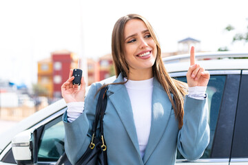 Young pretty woman holding car keys at outdoors intending to realizes the solution while lifting a finger up - obrazy, fototapety, plakaty