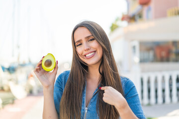 Young woman holding an avocado at outdoors with surprise facial expression