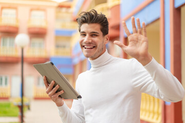 Young caucasian man holding a tablet at outdoors saluting with hand with happy expression