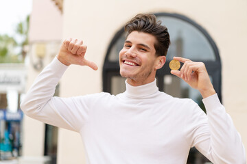 Young caucasian man holding a Bitcoin at outdoors proud and self-satisfied