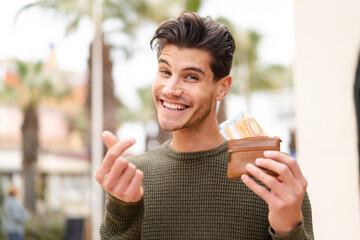 Young caucasian man at outdoors holding wallet with money with money gesture