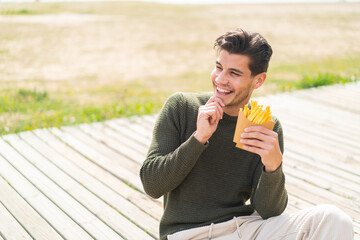Young caucasian man holding fried chips at outdoors thinking an idea and looking side