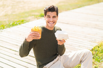 Young caucasian man at outdoors taking fried chips and takeaway box food with happy expression