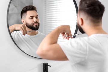 Handsome young man trimming beard with scissors near mirror in bathroom