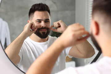 Handsome young man touching mustache near mirror in bathroom