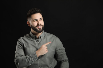 Young man with mustache pointing at something on black background. Space for text