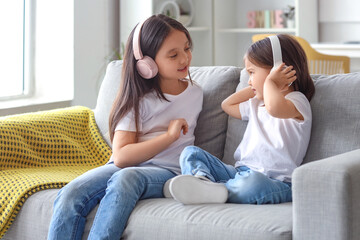 Cute little Asian happy sisters in headphones listening music on sofa at home