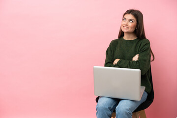 Young caucasian woman sitting on a chair with her laptop isolated on pink background looking up...