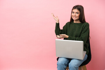 Young caucasian woman sitting on a chair with her laptop isolated on pink background extending...