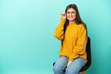 Young caucasian woman sitting on a chair isolated on blue background with glasses and happy