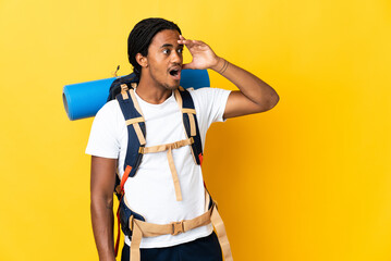 Young mountaineer man with braids with a big backpack isolated on yellow background doing surprise...