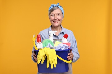 Happy housewife holding bucket with cleaning supplies on orange background