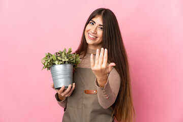 Young gardener girl holding a plant isolated on pink background inviting to come with hand. Happy...