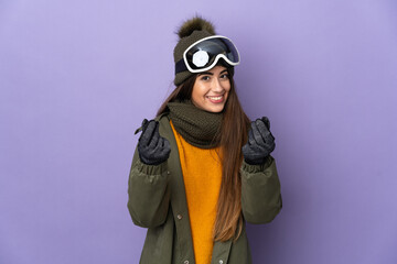 Skier caucasian girl with snowboarding glasses isolated on purple background making money gesture