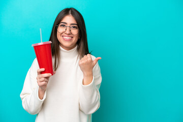 Young caucasian woman drinking soda isolated on blue background pointing to the side to present a...