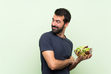 Young handsome man with salad over isolated green wall with arms crossed and happy