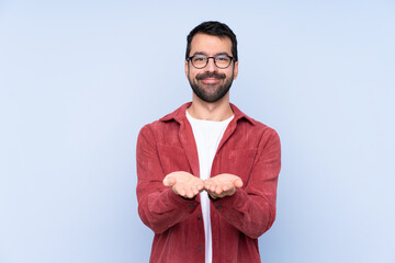 Young caucasian man wearing corduroy jacket over blue background holding copyspace imaginary on the palm to insert an ad