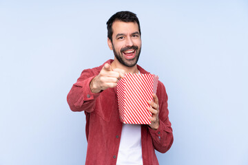 Young caucasian man over blue background holding a big bucket of popcorns while pointing front