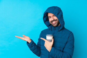 Man wearing winter jacket and holding a takeaway coffee over isolated blue background holding copyspace imaginary on the palm to insert an ad