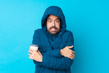 Man wearing winter jacket and holding a takeaway coffee over isolated blue background freezing