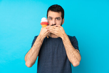 Young man with a cornet ice cream over isolated blue background covering mouth with hands
