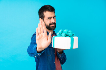 Young handsome man with a big cake over isolated blue background making stop gesture and...
