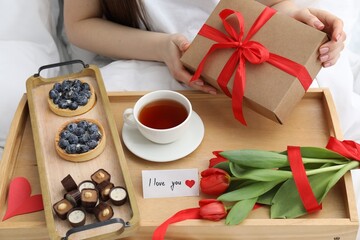 Tasty breakfast served in bed. Woman with gift box, desserts, tea, flowers and I Love You card at home, closeup
