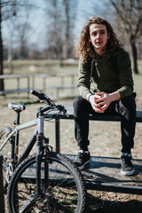 A curly-haired young man enjoys a break on a park bench beside his mountain bike, reflecting a moment of relaxation outdoors.