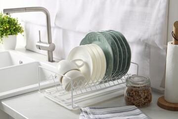 Drainer with different clean dishware and cups on white table in kitchen