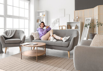 Smiling young woman with air conditioner remote control lying on sofa in living room