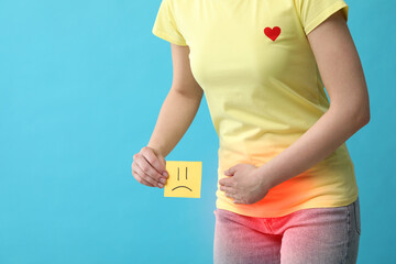 Woman holding sticky note with drawn sad face and suffering from cystitis symptoms on light blue...