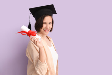 Happy female student in graduation hat with diploma winking on lilac background