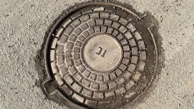 A iron hatch with a pattern above an observation well is located on auto road in daytime. Close-up