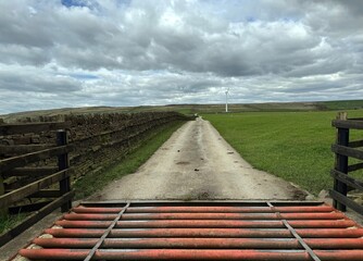 A rural lane with dry stone walls meanders toward a far-off wind turbine, set against a cloud-filled sky, with a cattle grid in the foreground near, Green Lane, Bradley, UK.