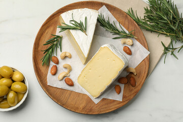 Plate with pieces of tasty camembert cheese, nuts, olives and rosemary on white marble table, flat lay