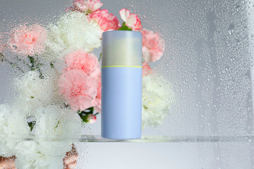 Bottle with moisturizing cream and beautiful flowers on light background, view through wet glass....
