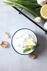 Delicious yogurt with dill in bowl, garlic, green onion and lemon on light textured table, flat lay