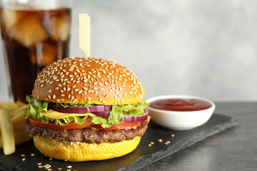 Burger with delicious patty, soda drink, french fries and sauce on dark table against gray background, closeup